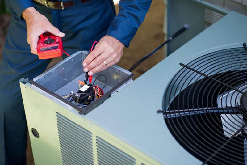 A technician using a multimeter to test the electrical connections inside the open control panel of an outdoor air-conditioning condenser unit.