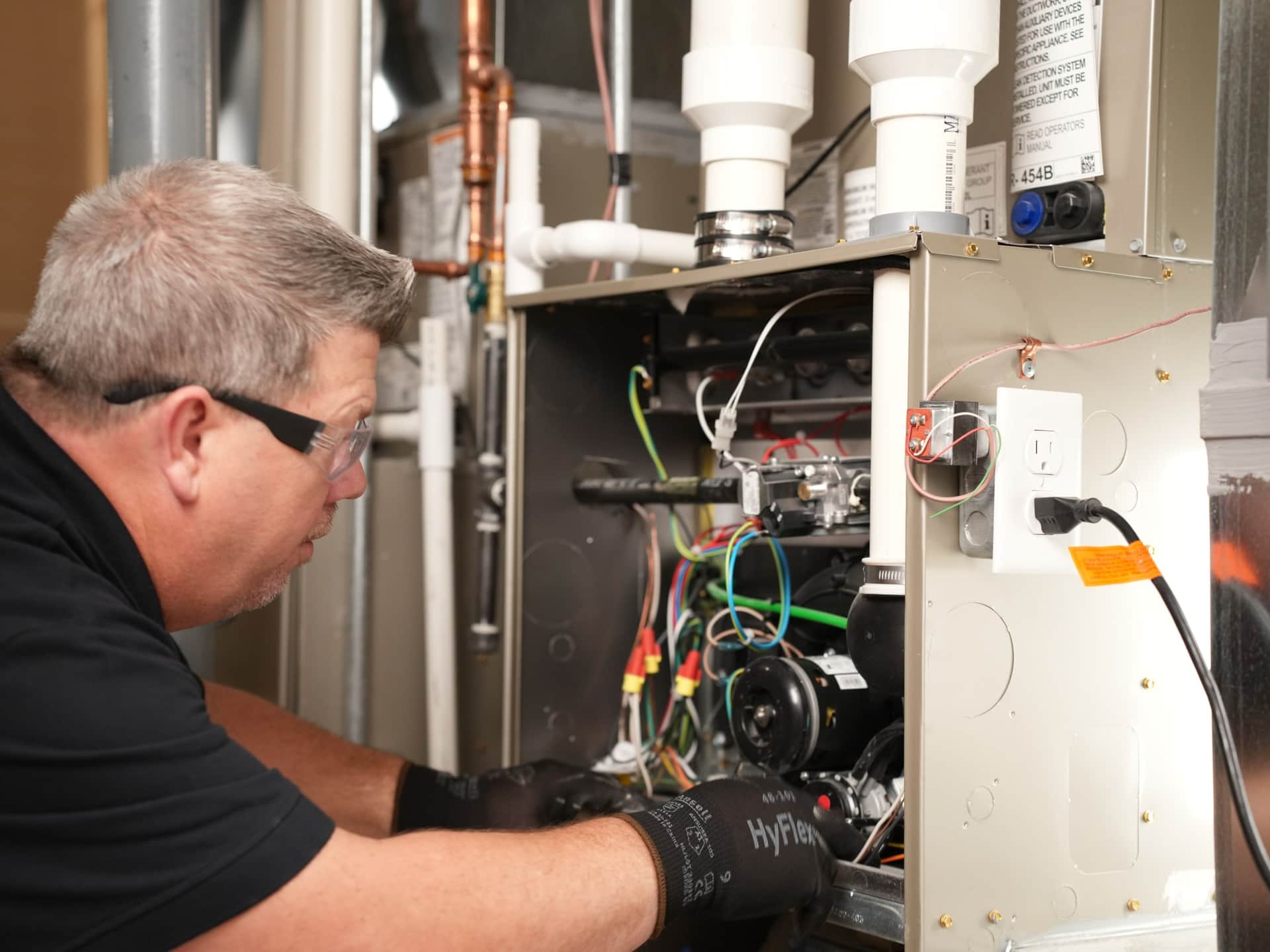 A technician in safety glasses and work gloves examining the open cabinet of a residential gas furnace, with the gas valve, blower and venting exposed.
