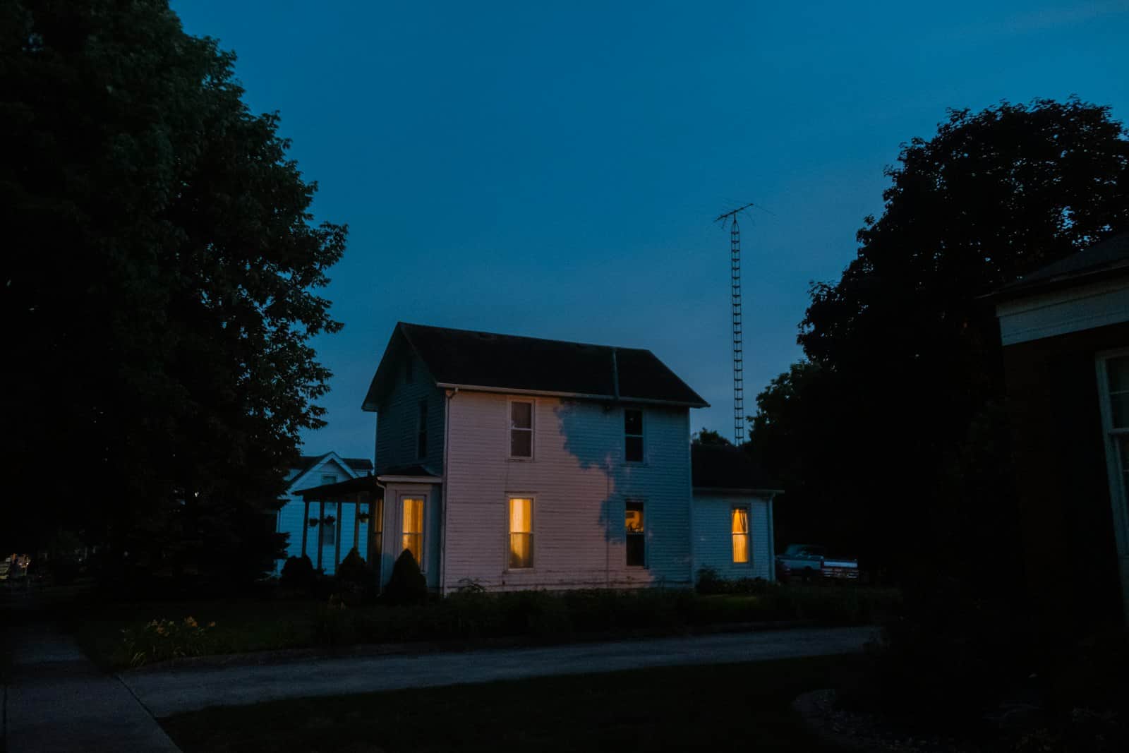An ordinary two-storey clapboard house at dusk with a few downstairs windows lit, a neighboring house and mature trees on a quiet residential street.