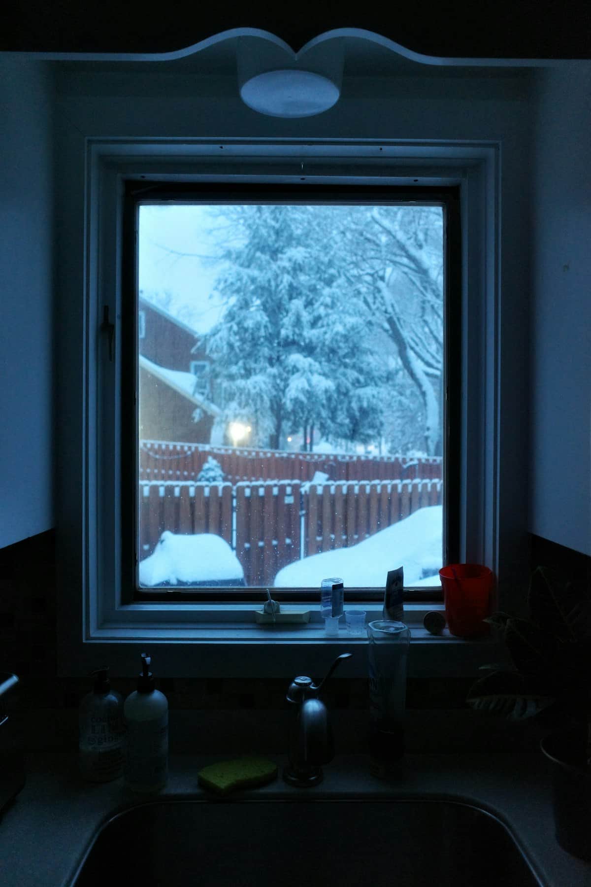 A darkened kitchen window looking out onto a backyard, fence and trees under deep snow.