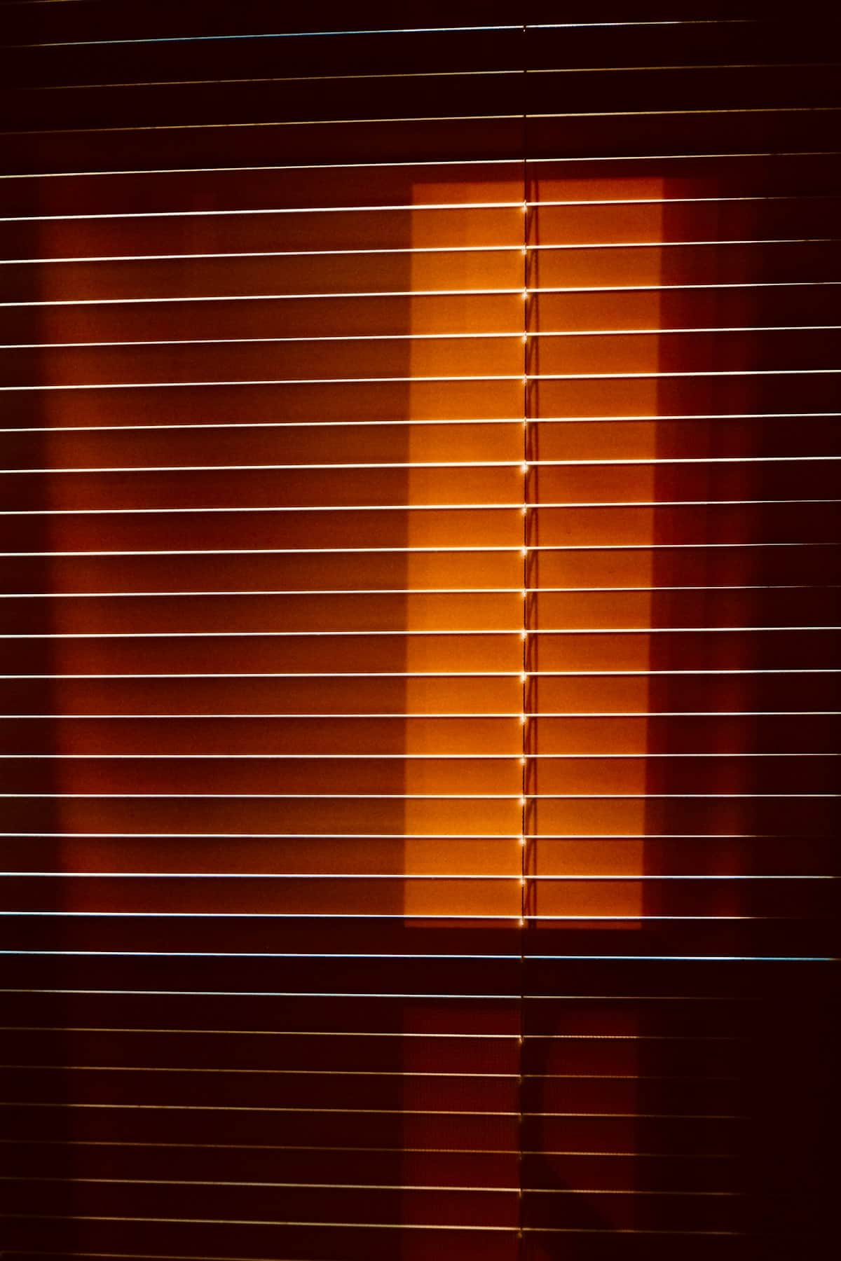 Hot afternoon sunlight burning through the slats of a closed window blind in a darkened room.